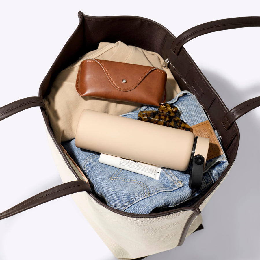 Open tote bag with folded clothes and a brown leather pouch on a light gray background